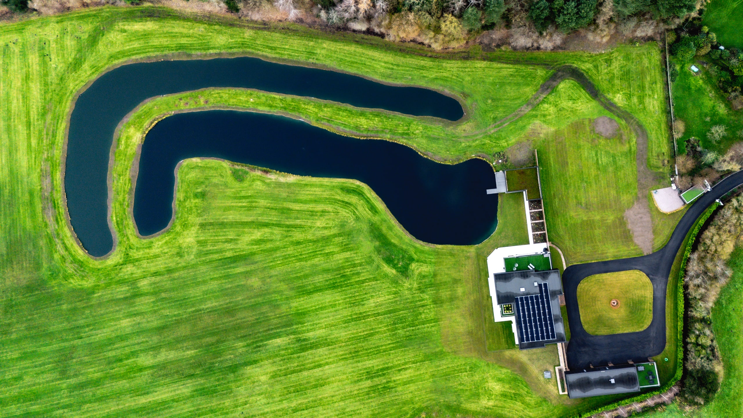 Aerial view of modern facility with curved pond, manicured green lawns, solar-paneled building, and baseball diamond